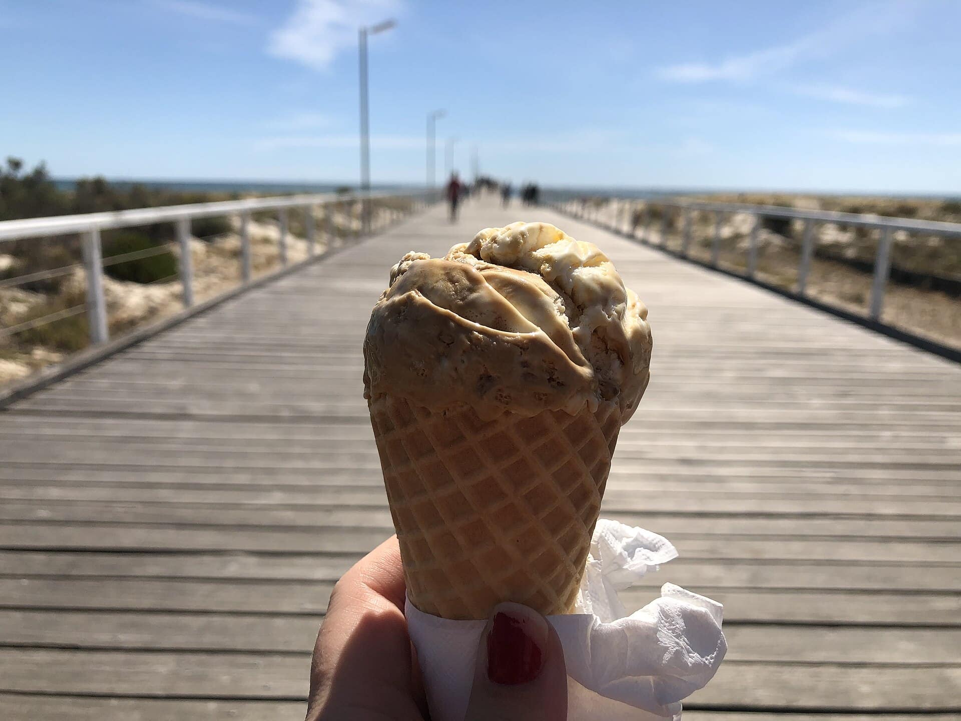 A hand holding a vanilla ice cream cone on a boardwalk by the beach