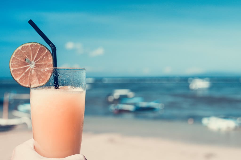 Pink tropical cocktail with a lime slice and straw on a table by the beach