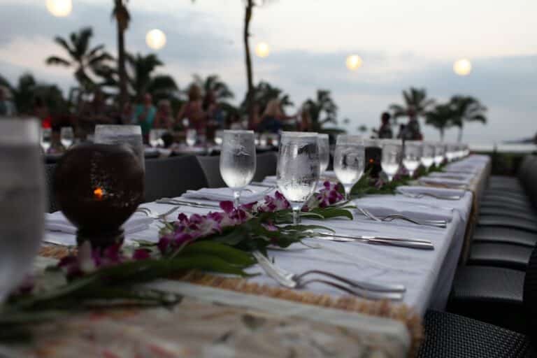 A long dinner table set with glassware and flowers on the beach at sunset